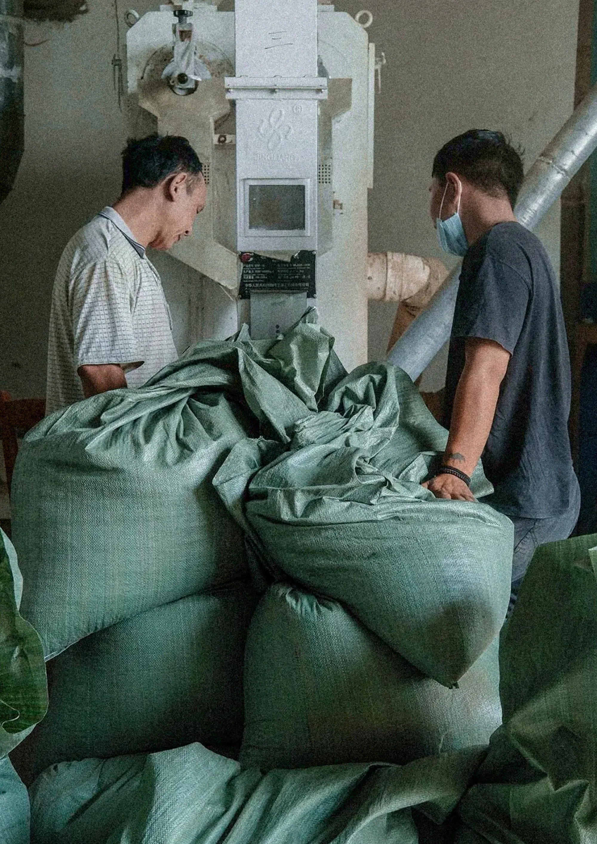 Two men beside stacked sacks of green coffee, in front of the hulling machine